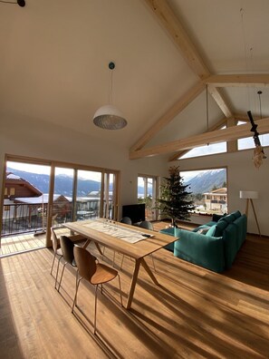 Amplia sala de estar con una mesa de comedor de madera, sillas y un sofá. Grandes ventanas ofrecen vistas a montañas y un balcón.