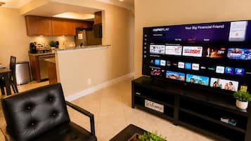 A modern living room with a flat-screen TV displaying a streaming service interface, a black leather sofa, a coffee table with a plant, and a kitchen area in the background.