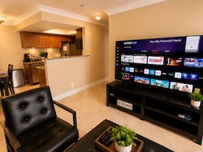 A modern living room with a flat-screen TV displaying a streaming service interface, a black leather sofa, a coffee table with a plant, and a kitchen area in the background.