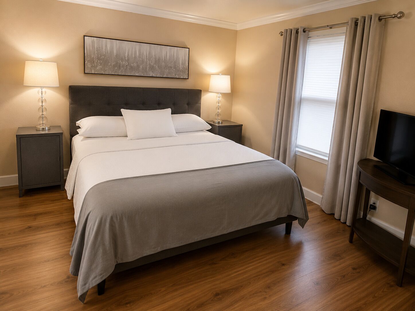 Un lit bien fait, recouvert d’une couvre-lit grise, deux tables de chevet avec des lampes, un téléviseur à écran plat posé sur un meuble et une fenêtre avec des rideaux.