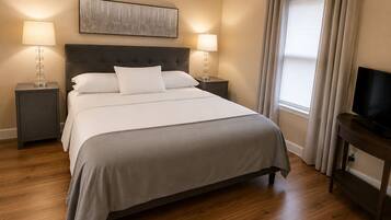 Un lit bien fait, recouvert d’une couvre-lit grise, deux tables de chevet avec des lampes, un téléviseur à écran plat posé sur un meuble et une fenêtre avec des rideaux.
