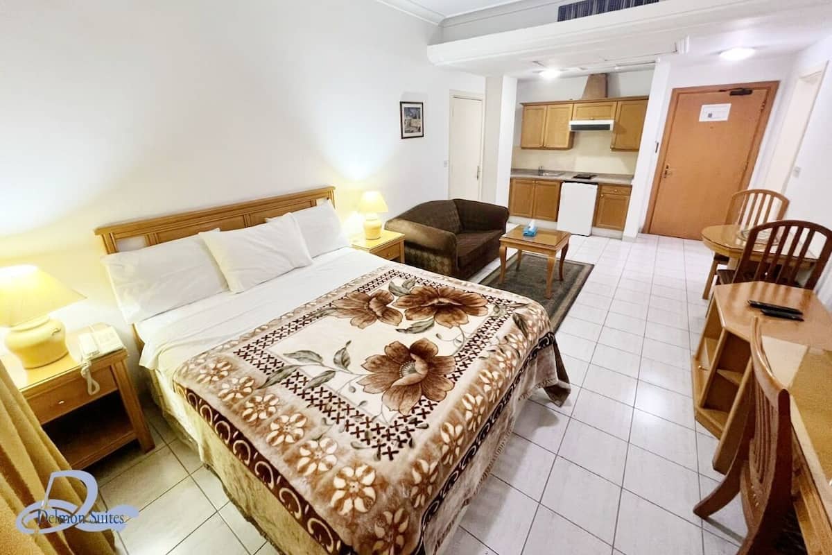 A hotel room with a bed, bedside tables, a desk, and a chair. There is a kitchenette in the background.