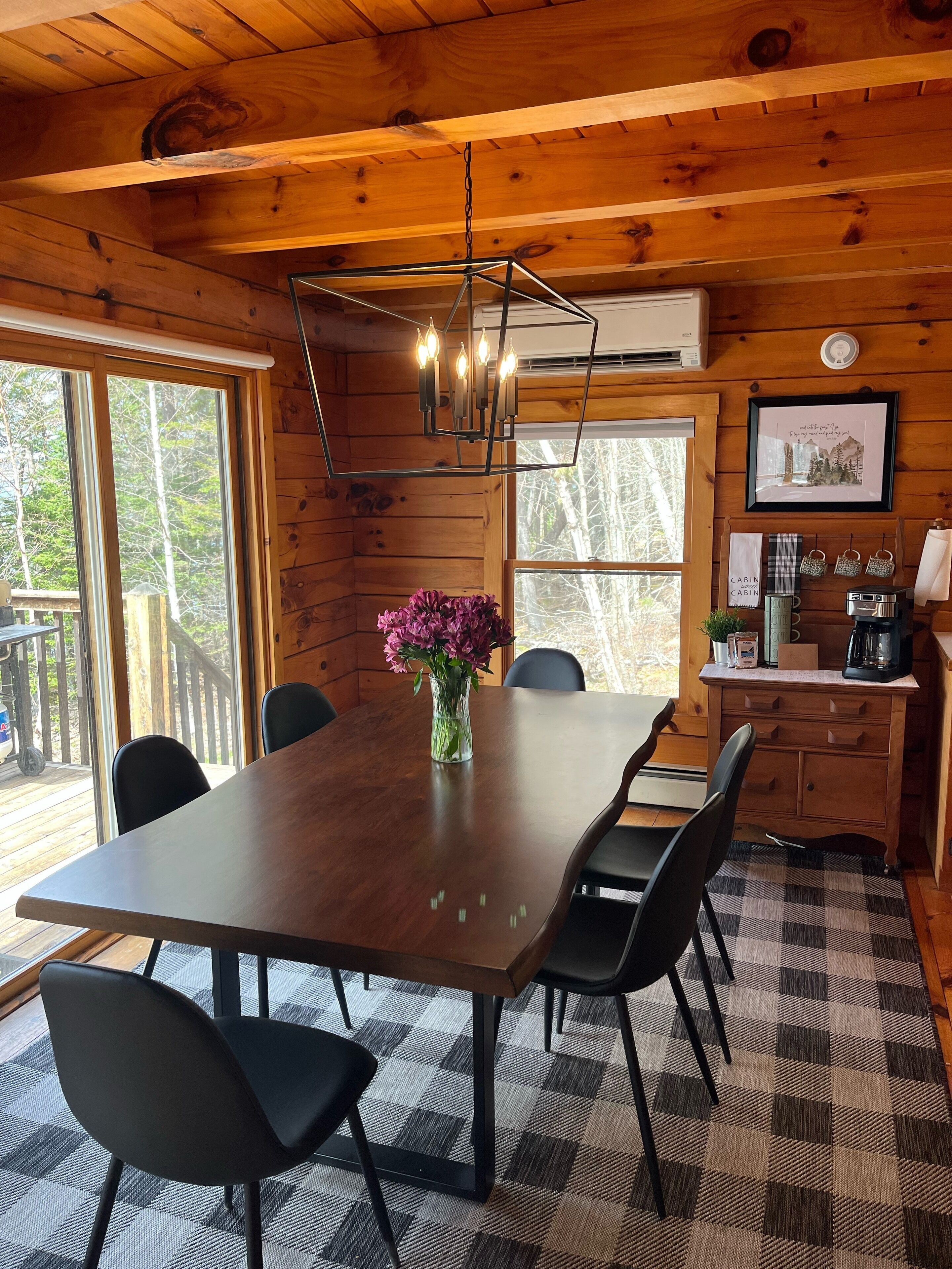 A wooden cabin interior with a dining table, chairs, a vase of flowers, a coffee maker, and a framed picture on the wall.