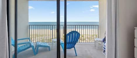 A balcony with a view of the beach, featuring blue chairs and a small table.