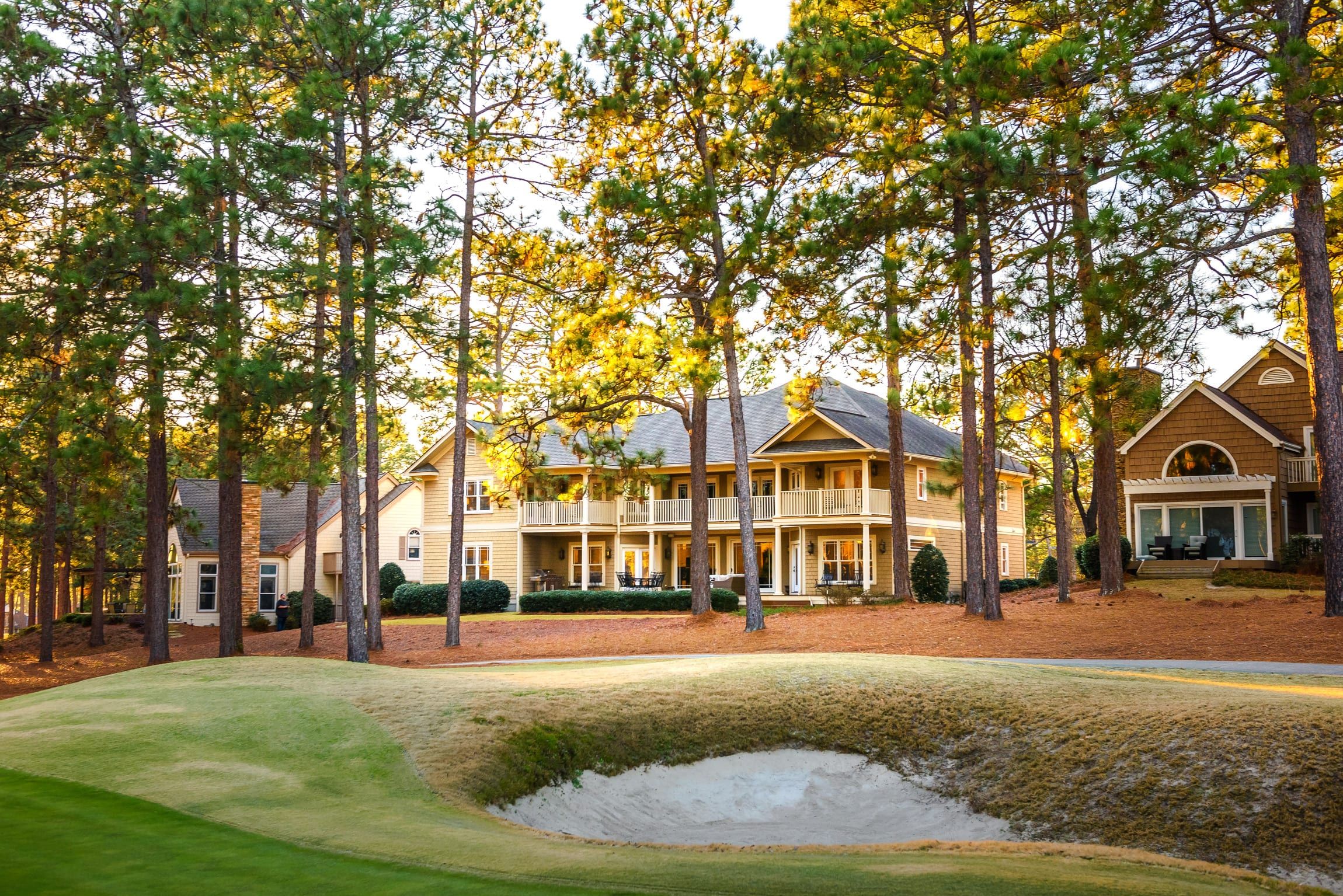 A golf course with a sand bunker, surrounded by tall trees and a large, multi-story building with a porch.