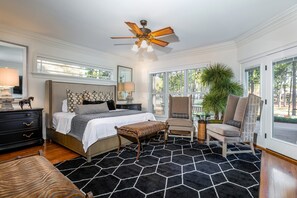 Ein Schlafzimmer mit einem Bett, einem Schreibtisch, einem Sessel, einer Sitzbank mit Zebra-Muster, einem Deckenventilator und einem großen Fenster mit Blick auf Bäume.