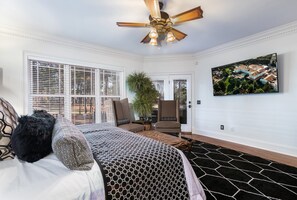 A bedroom with a bed, a ceiling fan, a large window with blinds, a potted plant, a chair, and a painting on the wall.