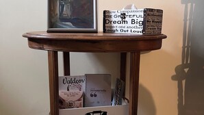 Une petite table en bois sur laquelle est posée une peinture encadrée, une boîte contenant des phrases inspirantes et un tiroir rempli de livres et d’un téléphone.