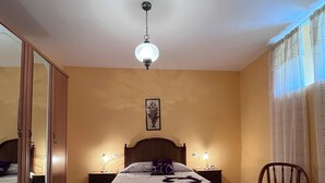 Une chambre à coucher comprenant un lit, des lampes de chevet, une chaise en bois, un banc, un luminaire suspendu et un miroir.