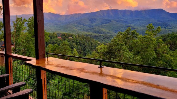 Una terraza de madera con barandilla metálica que da a un paisaje montañoso con bosque denso y algunos edificios a lo lejos.