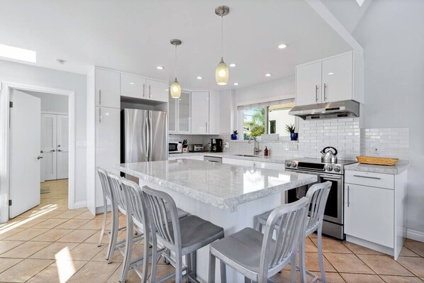 A modern kitchen with white cabinetry, a large island with bar stools, stainless steel appliances, and pendant lighting. - Mission Beach Steps to Beach Remodeled Home with AC, fireplace, and Parking (San Diego)