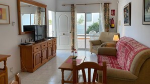 Ein Wohnzimmer mit Fernseher, Holzmöbeln, einer Couch, einem Tisch und Blick durch eine Glastür auf einen Balkon.