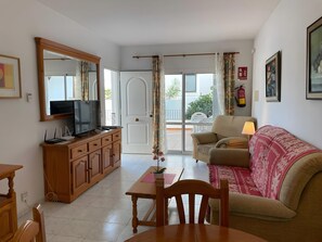 Un soggiorno con televisione, mobili in legno, un divano, un tavolo e vista su un balcone esterno attraverso una porta di vetro.