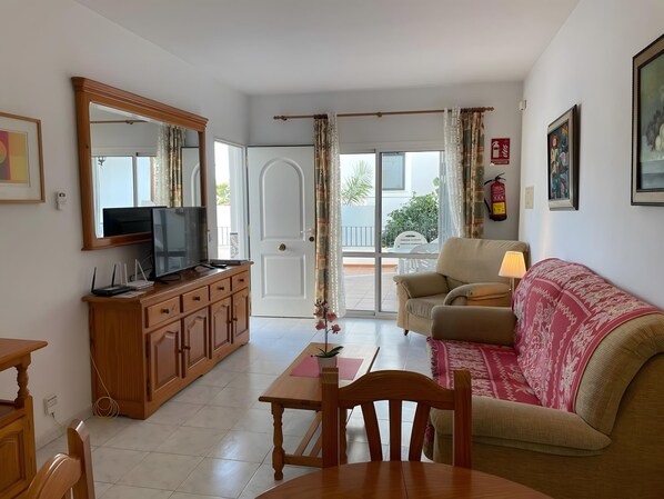 A living room with a television, wooden furniture, a sofa, a table, and a view through a glass door to an outdoor balcony.