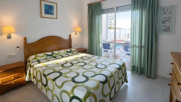 Ein Schlafzimmer mit einem Bett, zwei Nachttischlampen, einer Holzkommode, einem Fenster mit Vorhängen und Blick nach draußen.
