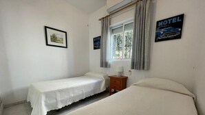Ein Zimmer mit zwei Betten, einem Nachttisch, einem Fenster mit Vorhängen und einem gerahmten Bild an der Wand.