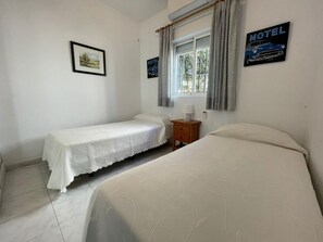 Una stanza con due letti, un comodino, una finestra con tende e un quadro appeso al muro.