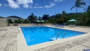 A rectangular swimming pool with a tiled deck, surrounded by lounge chairs and umbrellas, set against a backdrop of palm trees and a mountain. - Crystal Cove At Sapphire Beach (St Thomas)