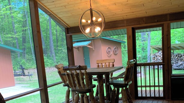 A wooden deck with a round table and chairs, a hanging light fixture, and a view of a cabin and forest. - Modern Cabin located between Grindstone and Lac Courte Oreilles (Hayward)
