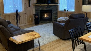 A cozy living room with a fireplace, wooden walls, and a large window. - Modern Cabin located between Grindstone and Lac Courte Oreilles (Hayward)
