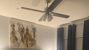 Un llit ben fet amb capçal, un ventilador de sostre, un quadre a la paret i una finestra amb cortines.
