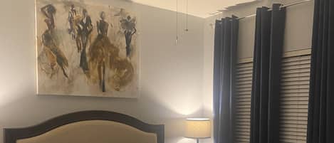 A neatly made bed with a headboard, a ceiling fan, a painting on the wall, and a window with curtains.