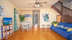 Un vaste espace de vie comprenant une table à manger, un canapé bleu, un ventilateur de plafond et un escalier menant à un autre niveau.