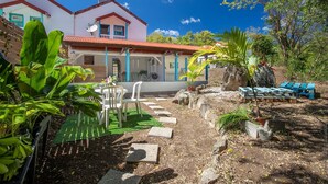 Une maison à deux étages avec un toit rouge et des murs blancs, une terrasse avec des chaises et un jardin où poussent différentes espèces de plantes.