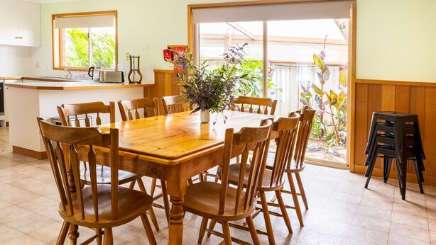 Comedor con mesa y sillas de madera, pequeño espacio de cocina con un lavamanos y una radio, y vistas a plantas a través de una ventana.