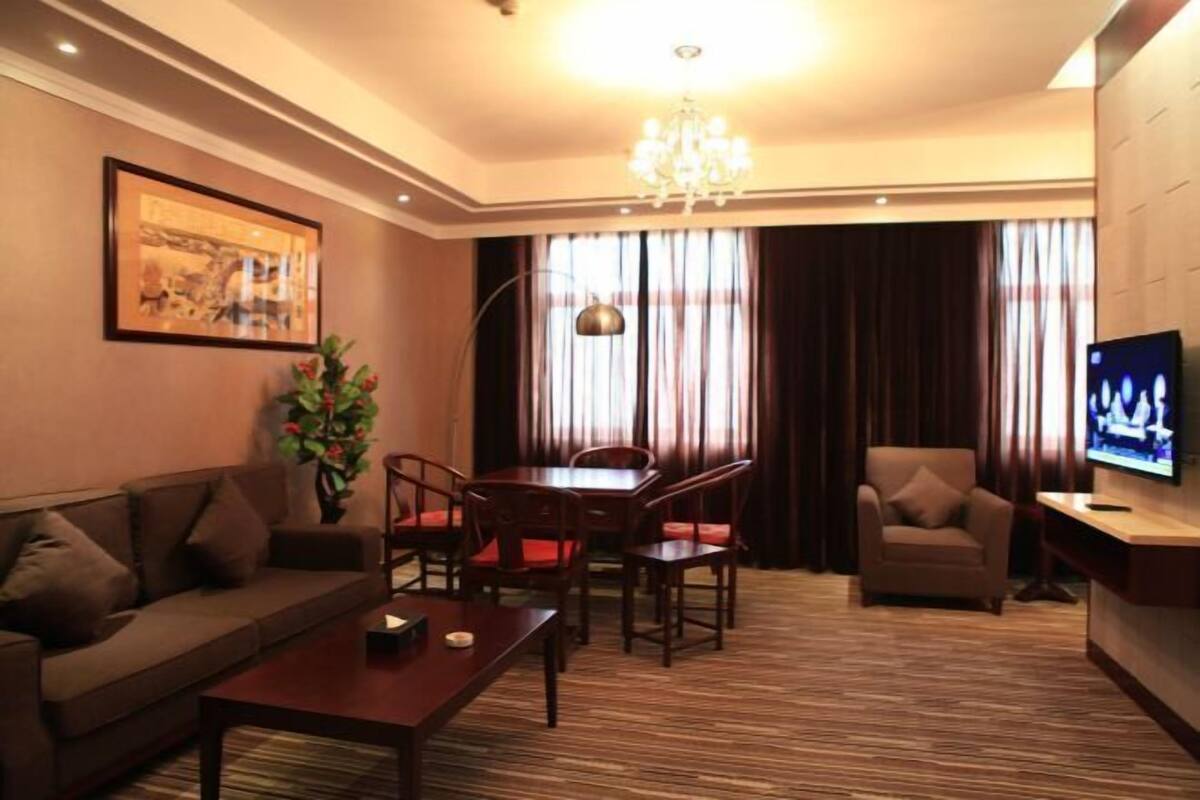 A hotel room with a sofa, dining table, chairs, a TV, a chandelier, and a painting on the wall.