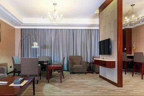 A hotel room with a flat-screen TV, a dining area with a table and chairs, a sofa, a small wooden side table, and a large window with curtains. - Jiutong Haiyuan International Hotel (Wuhan)