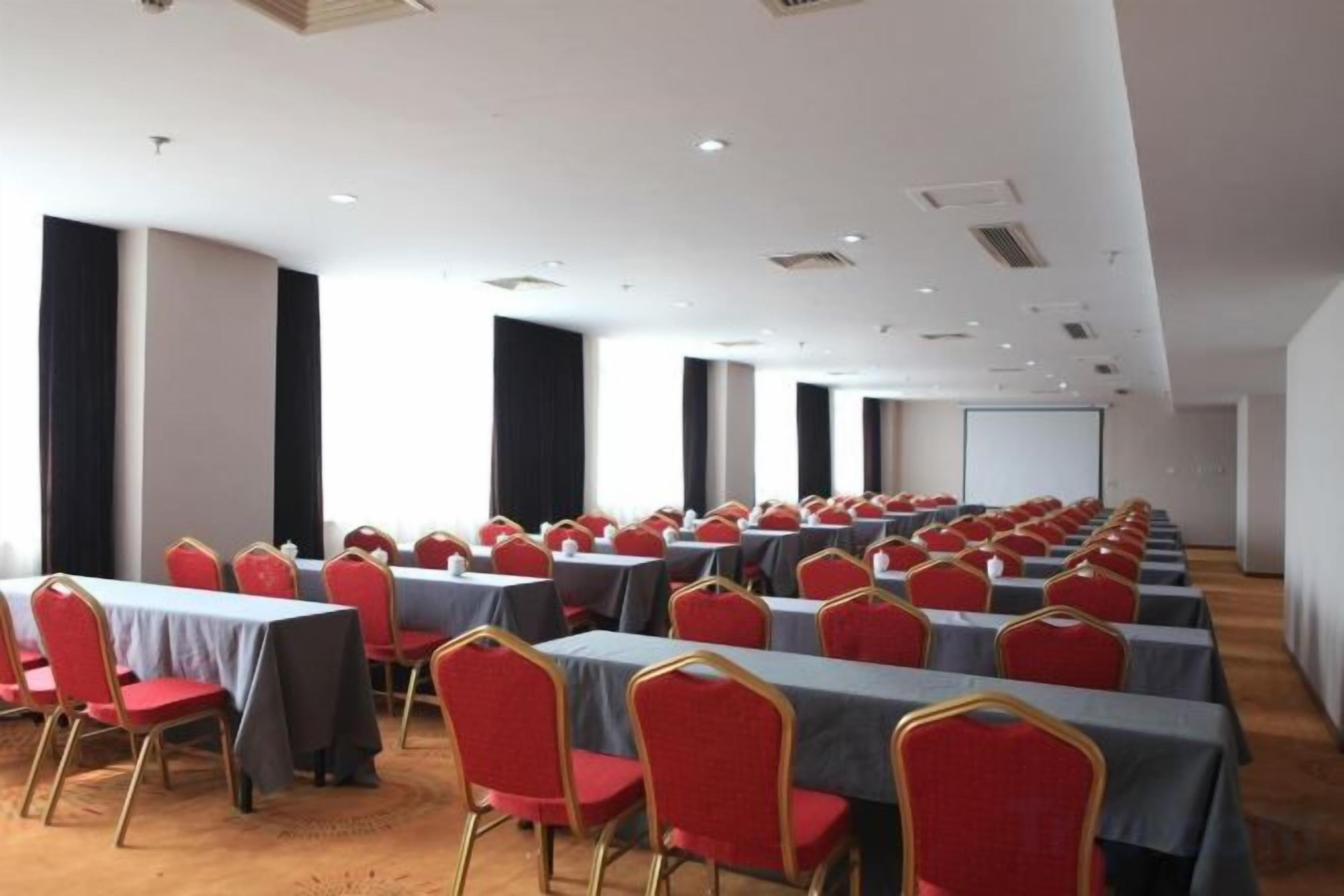 A conference room with rows of tables and chairs, a projector screen, and a carpeted floor.