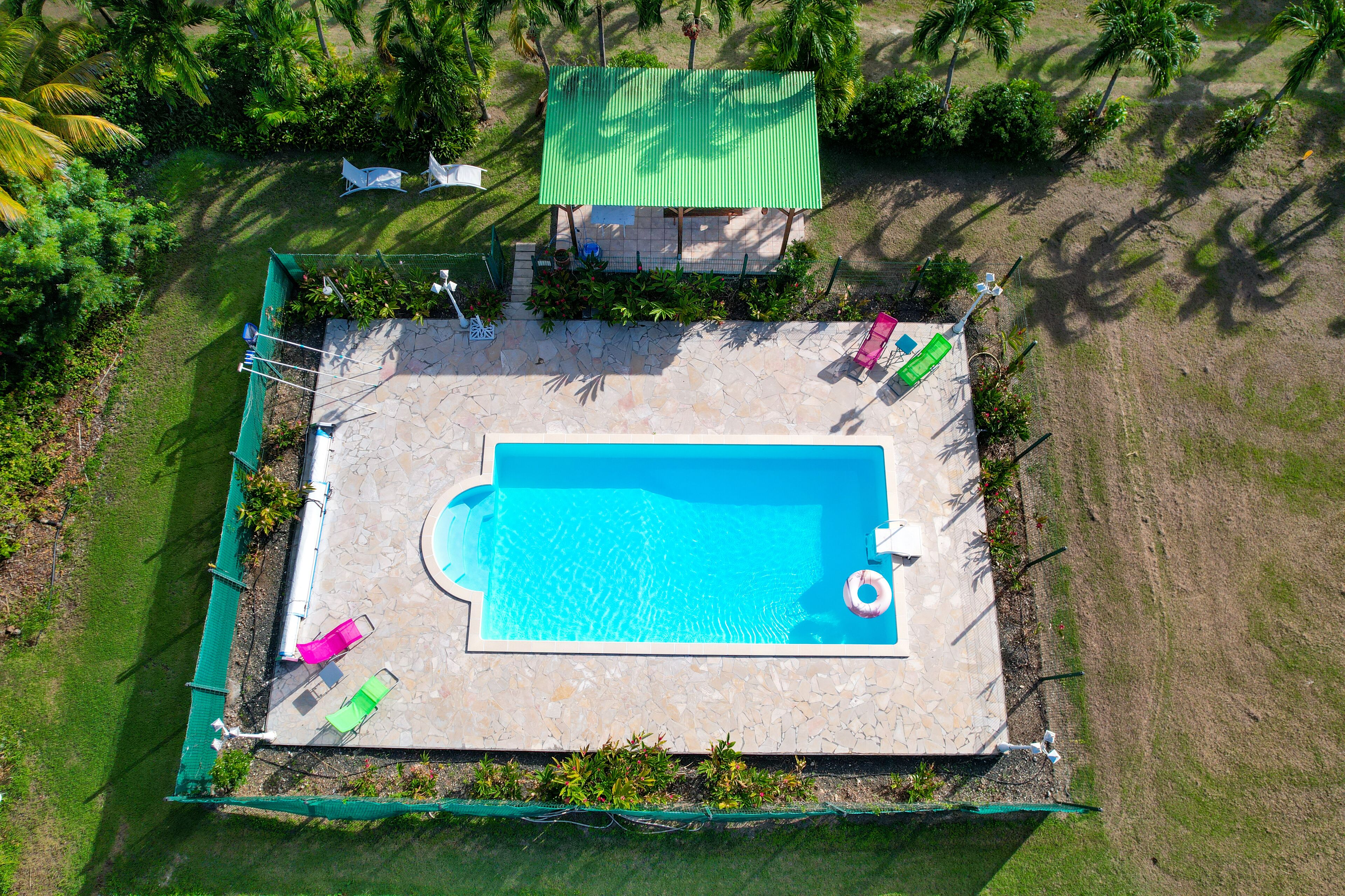 An aerial view of a swimming pool surrounded by a fenced area with a stone deck, a covered seating area, and lush greenery.