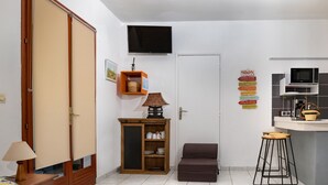 A compact living space with a kitchenette, a television mounted on the wall, a cabinet with kitchenware, and a small sofa. - Gîtes Papillon - Studio with swimming pool (Saint-François)