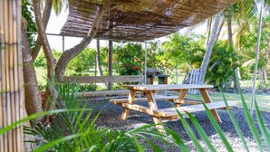 A wooden picnic table and benches under a thatched roof, surrounded by gravel and greenery. - Gîtes Papillon - Studio with swimming pool (Saint-François)