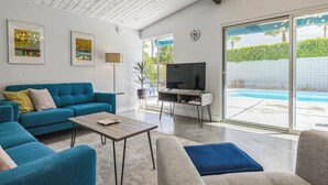A modern living room with a blue sofa, a coffee table, and a TV on a stand. There is a sliding glass door leading to a pool area. - Quintessential Midcentury Modern Alexander House (Palm Springs)