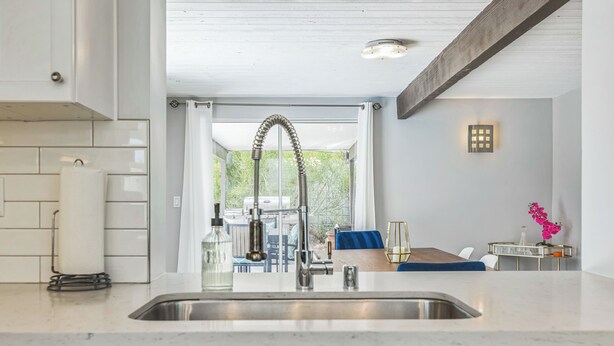 A modern kitchen with a stainless steel sink, white tiled backsplash, and blue cabinets. There is a dining area with a table and chairs visible through an open doorway. - Quintessential Midcentury Modern Alexander House (Palm Springs)