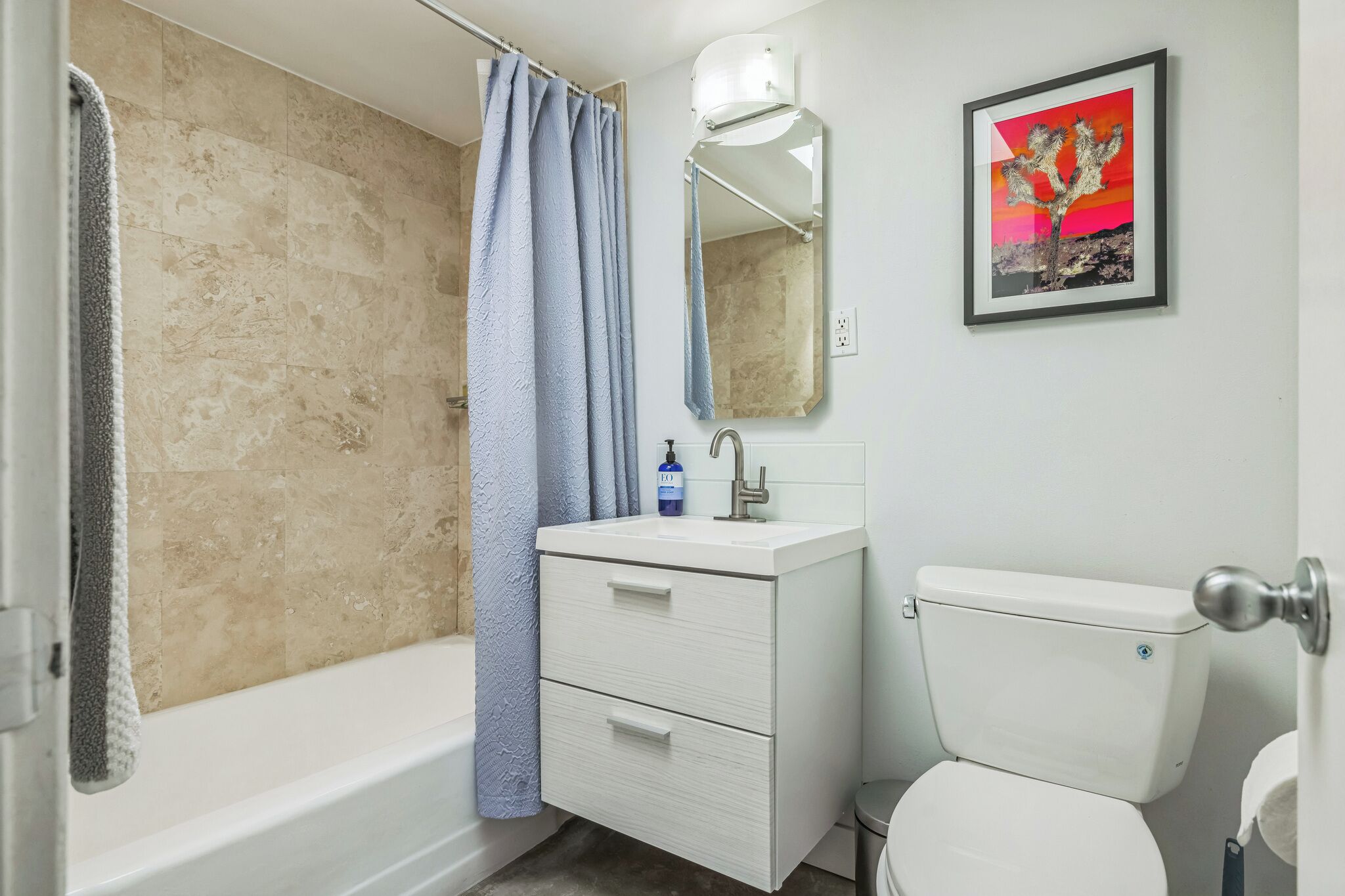 A bathroom with a bathtub, shower curtain, sink, mirror, toilet, and a framed picture on the wall.