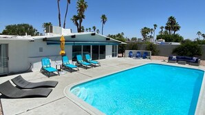 A rectangular swimming pool with clear blue water, surrounded by lounge chairs and a covered area with yellow umbrellas. - Quintessential Midcentury Modern Alexander House (Palm Springs)