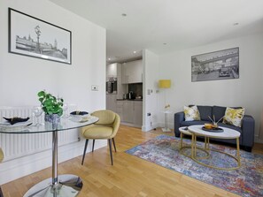 A modern living room with a glass dining table, a sofa, a coffee table, and a kitchen in the background.