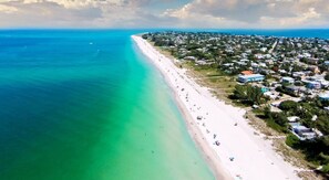 An aerial view of a coastal area with a long sandy beach, a clear blue ocean, and a densely populated residential area with houses and greenery. - STUNNING! Game Room, Pool, Beaches & Full Remodel! (Bradenton)