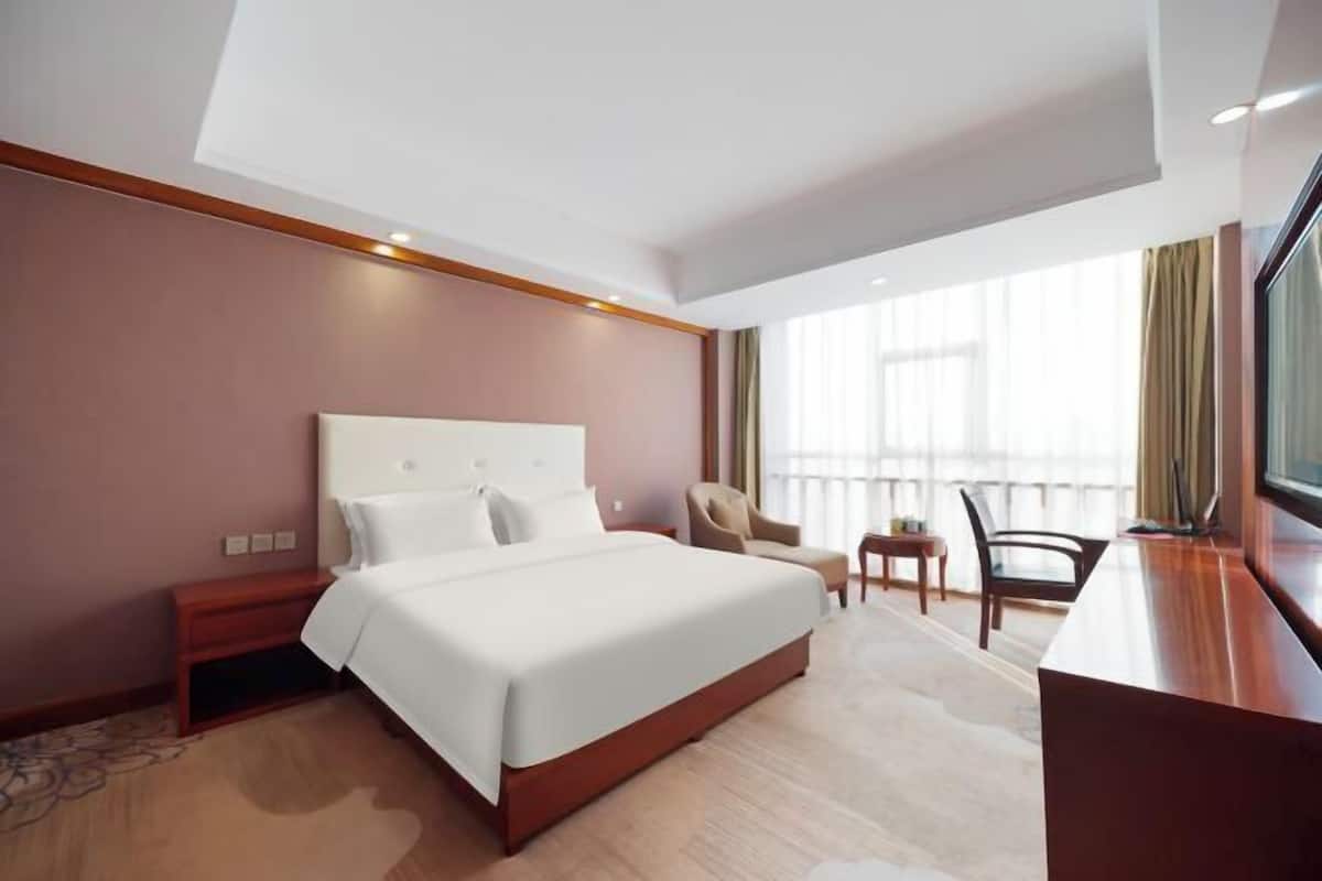A hotel room with a large bed, a desk, two chairs, and a window with curtains.