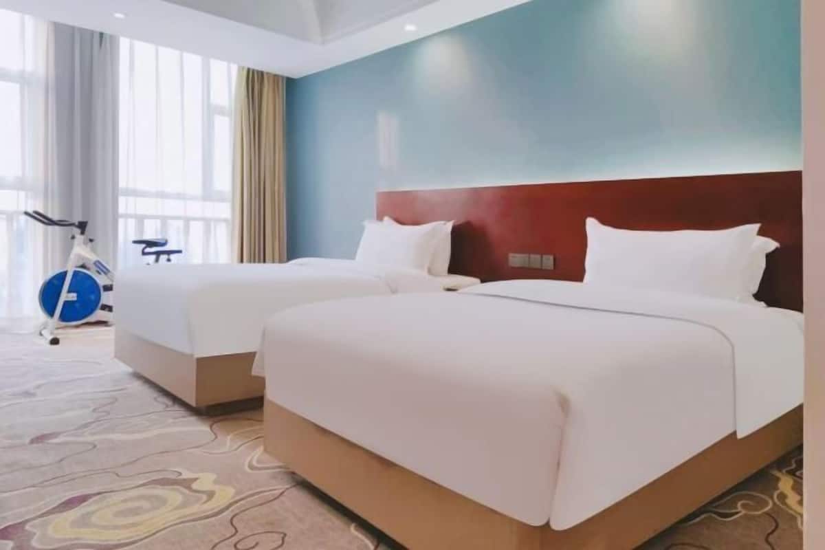 A hotel room with two beds, a workout bike, and a large window with curtains.