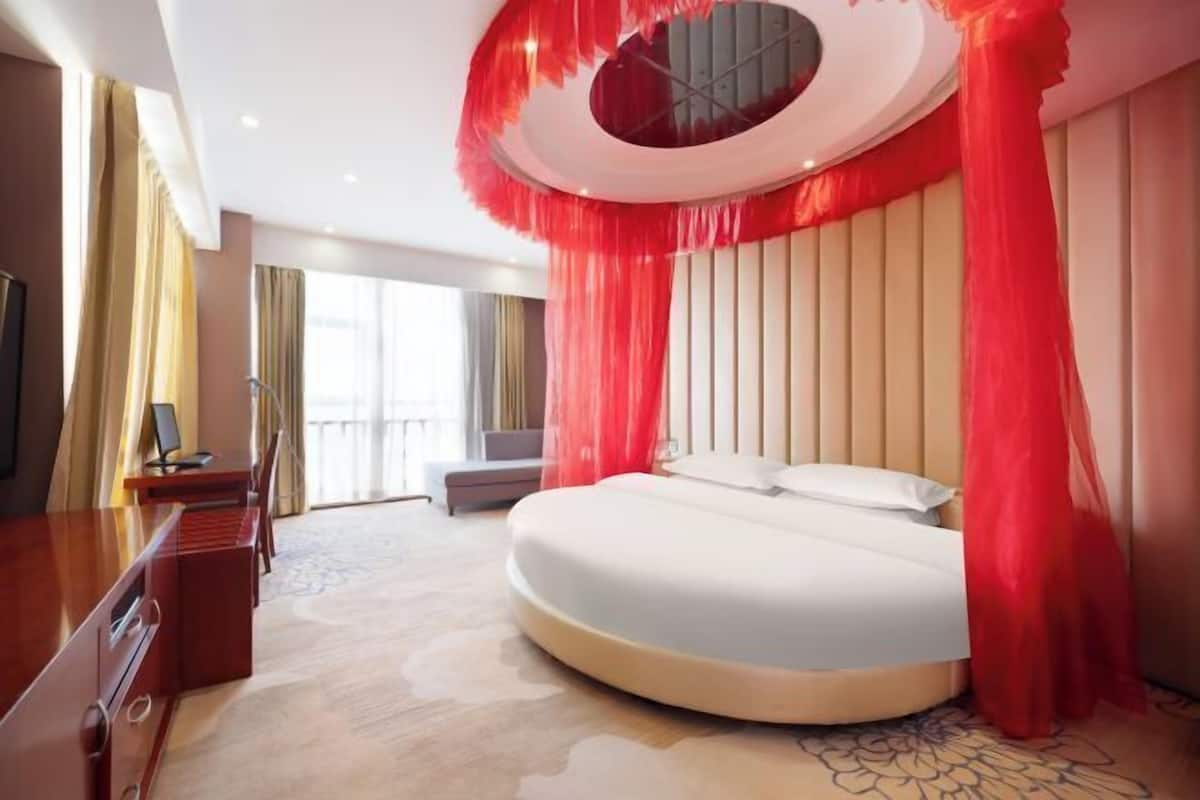A modern hotel room with a round bed, a desk, and a large window.