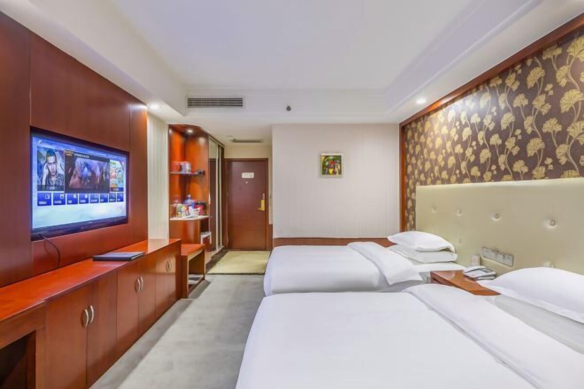 A hotel room with two beds, a TV on a wooden cabinet, a small shelf with items, and a floral-patterned wall.