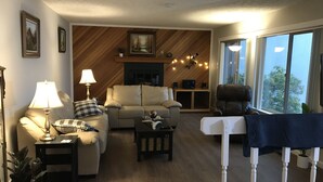 Una sala de estar acogedora con un sofá, sillones, una mesa de centro y una chimenea. Hay cuadros enmarcados en la pared, una lámpara y una planta.