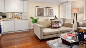 A modern living room with a beige sofa, a coffee table, and a floor lamp. There is a kitchen area with white cabinets and a sink. Two framed artworks are displayed on the wall. - Chancery Lane, Central London by Interhome (London City)