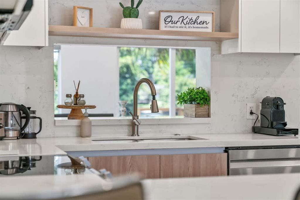 A modern kitchen with a stainless steel sink, wooden cabinets, a coffee maker, and a window with a view of greenery.