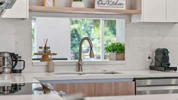 A modern kitchen with a stainless steel sink, wooden cabinets, a coffee maker, and a window with a view of greenery.