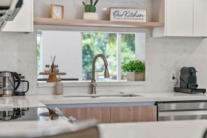 A modern kitchen with a stainless steel sink, wooden cabinets, a coffee maker, and a window with a view of greenery.